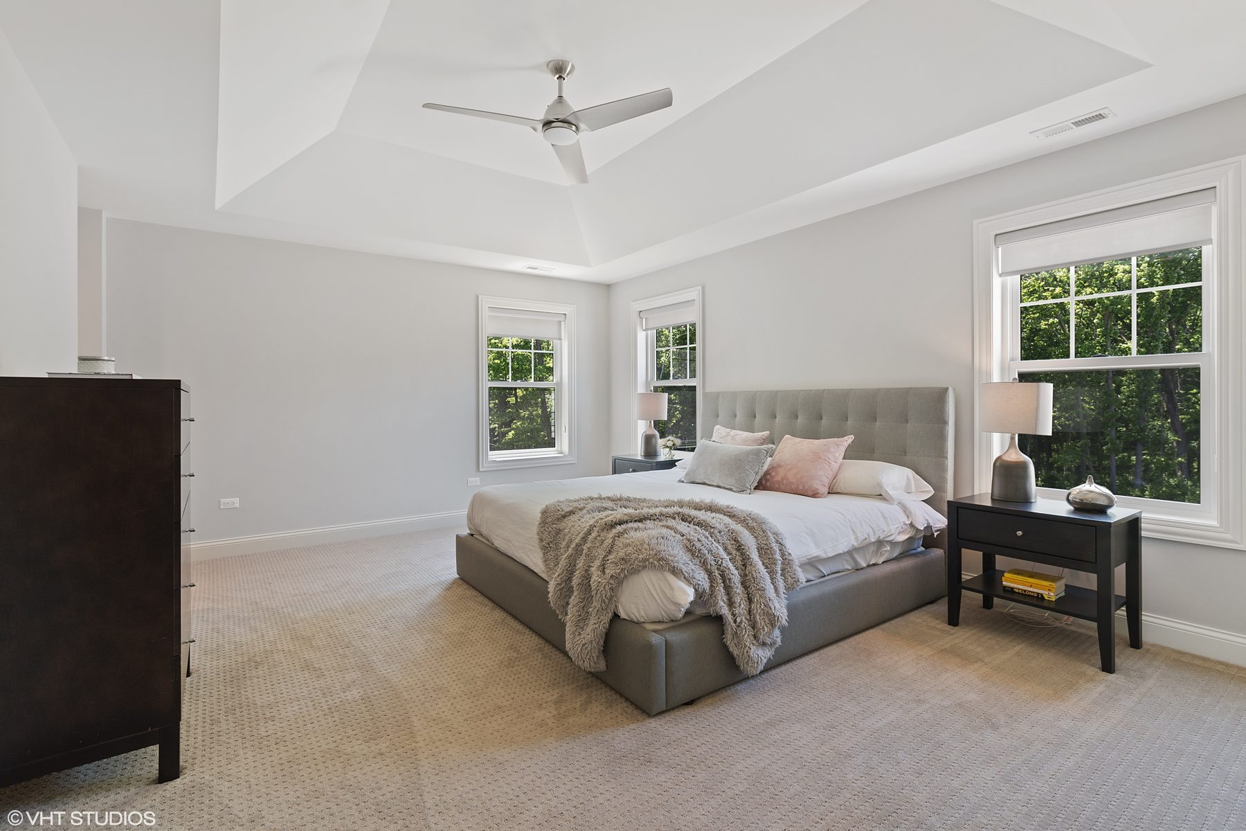 Primary bedroom with a tray ceiling, upholstered gray bed and windows onto the trees
